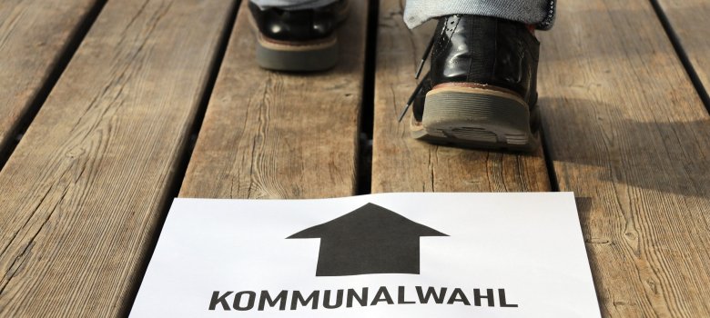 Zettel mit Aufschrift Kommunalwahl, schwarzer Pfeil geradeaus, im Hintergrund eine gehende Person auf Holzdielen