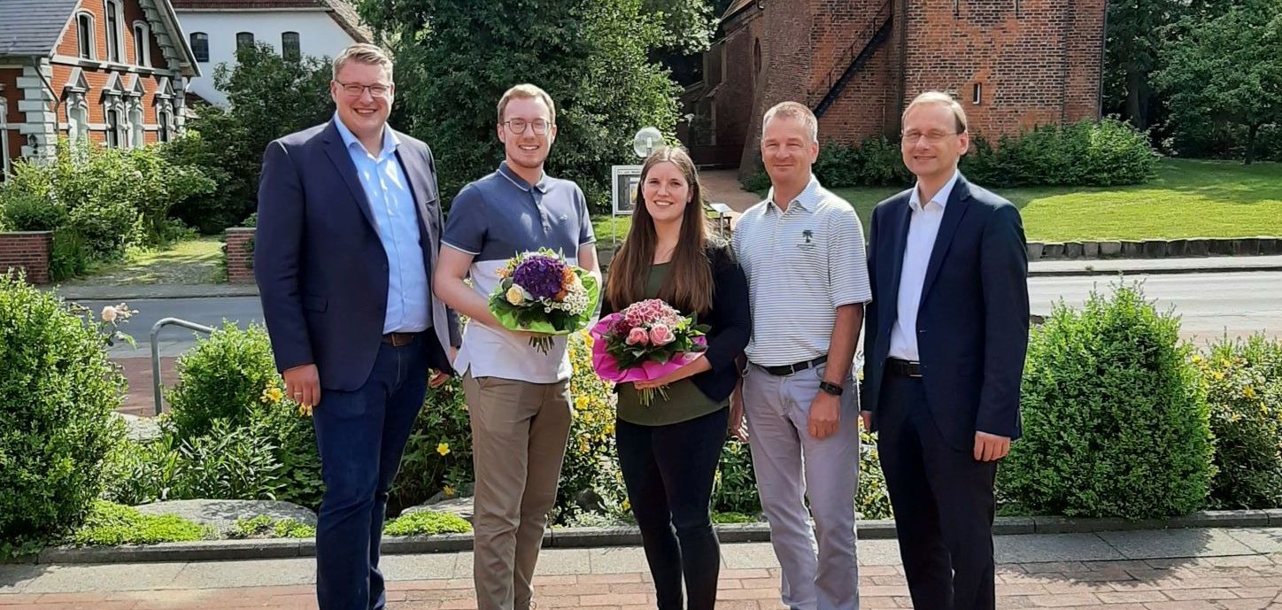 Mitarbeiter der Gemeinde stehen mit Blumenstrauß vor dem Rathaus. Im Hintergrund die Kirche. 